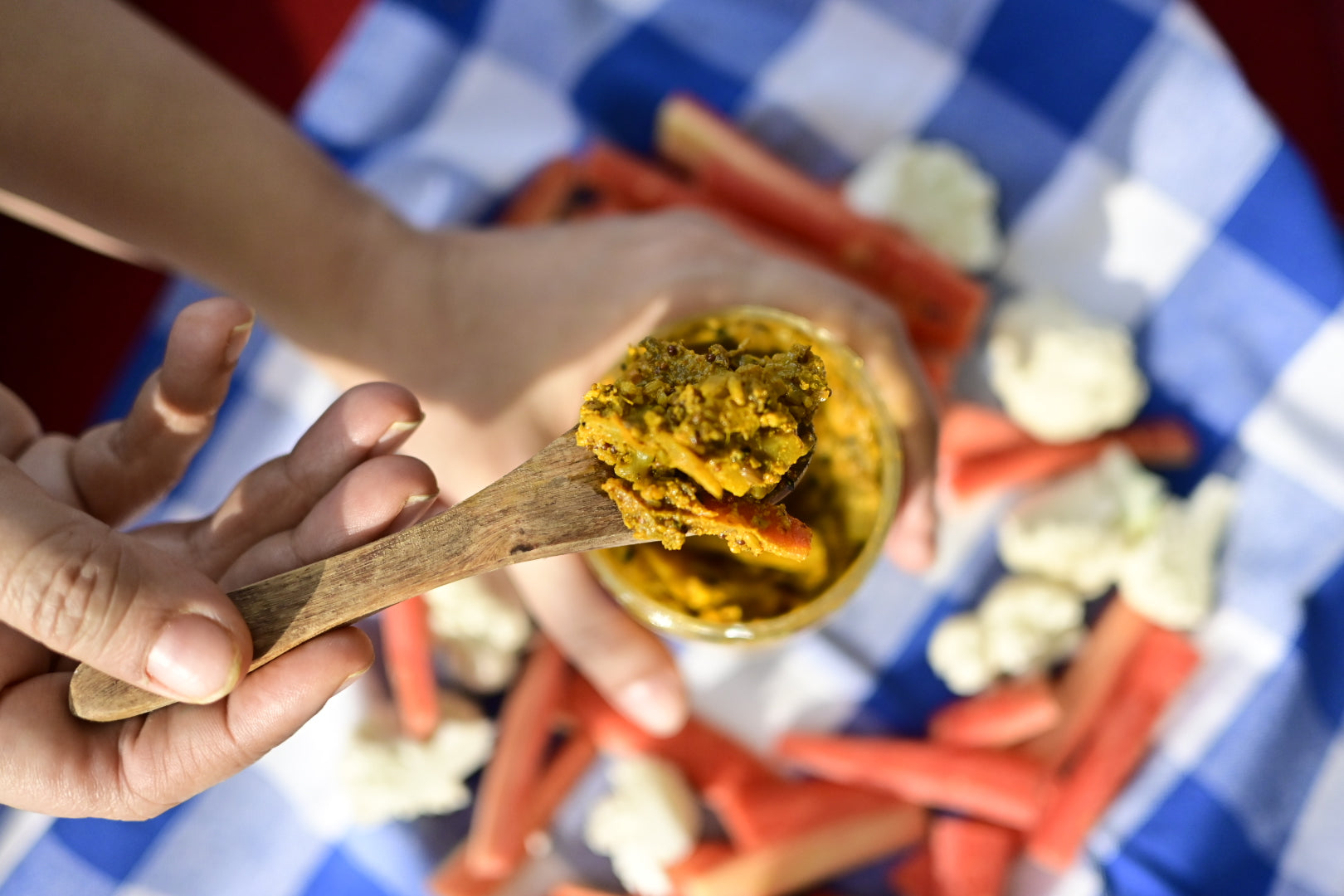 Hand holding a wooden spoon with Mix Veg Achaar over a container, surrounded by carrots and cauliflower on a checkered tablecloth.