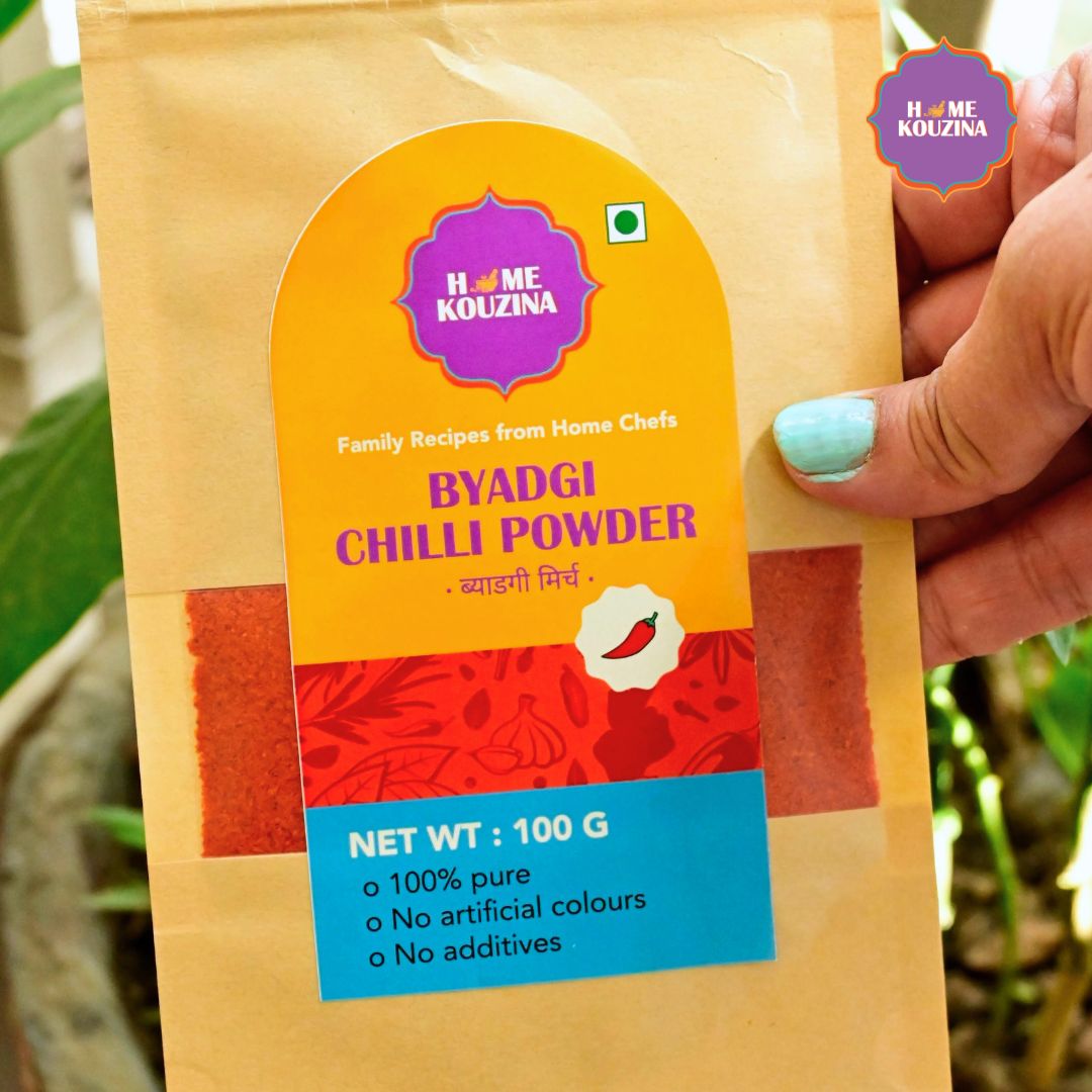 Hand holding Home Kouzina Byadgi Chilli Powder 100g pouch outdoors – premium-quality, slow-ground chilli powder with deep red color and low pungency
