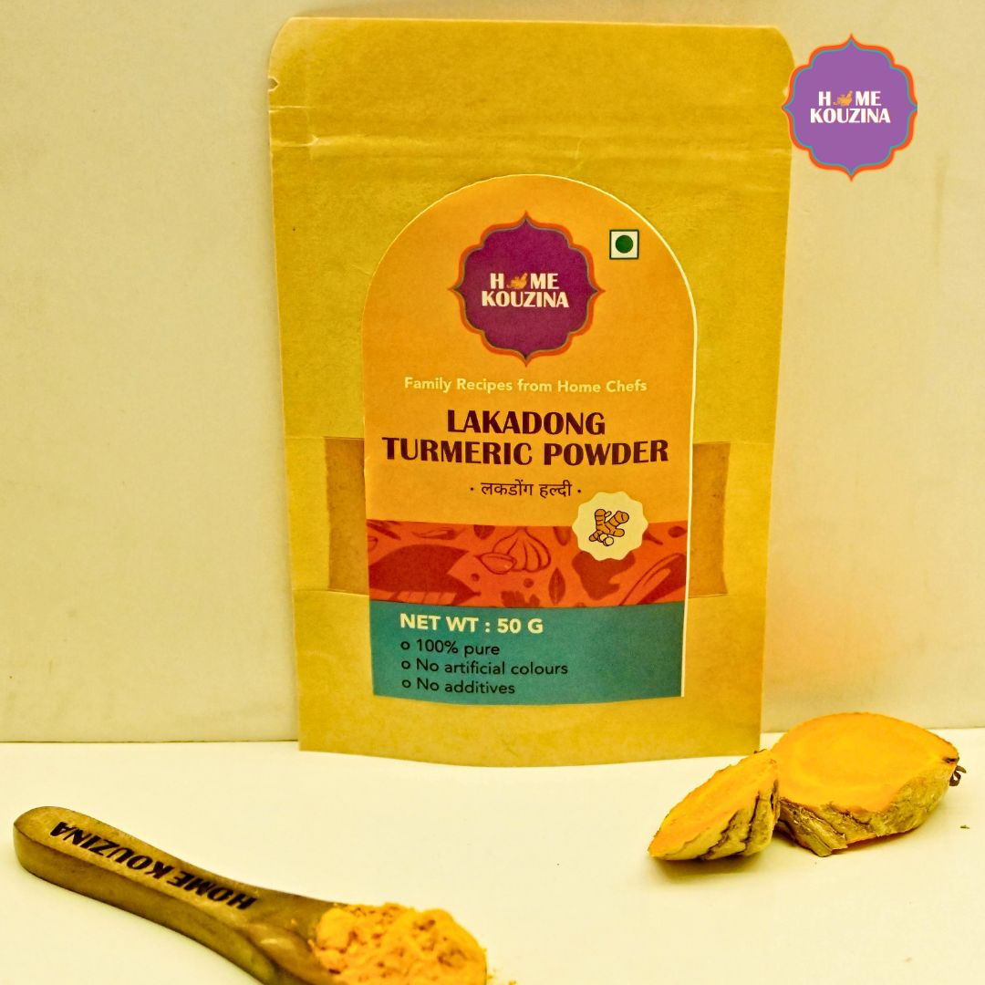 Front view of Home Kouzina Lakadong Turmeric Powder 50g pack on white background with spoonful of turmeric powder and whole turmeric roots – single-origin, high-curcumin spice from Meghalaya