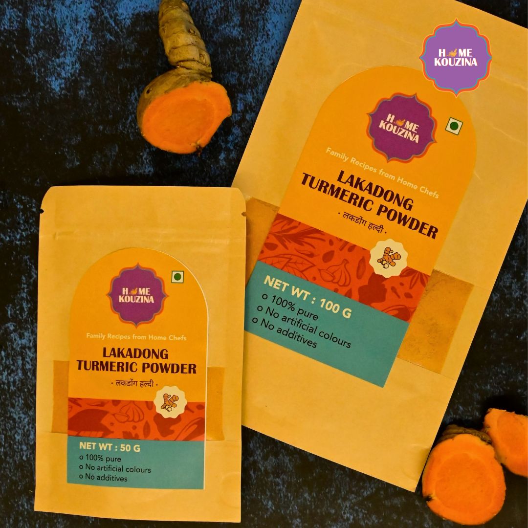 Front view of Home Kouzina Lakadong Turmeric Powder 50g and 100g pack on table with whole turmeric roots – single-origin, high-curcumin spice from Meghalaya