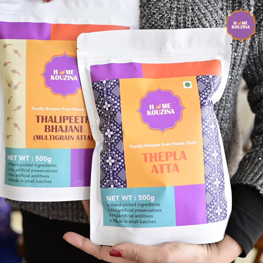 Two packages of 'Thepla Atta' and 'Thalipeeth Bhajani' from Home Kouzina held by a person.