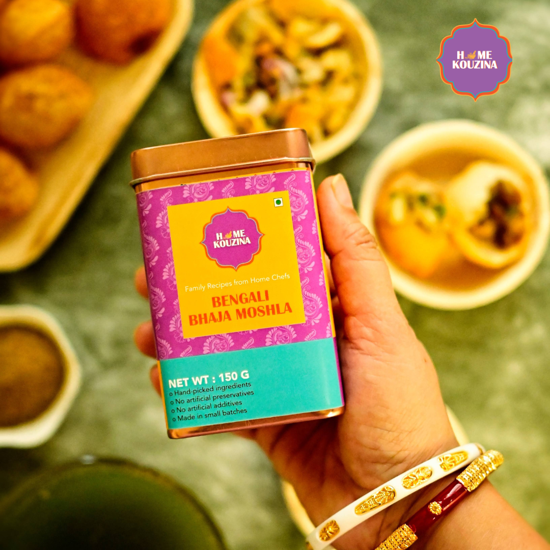 Top view of Home Kouzina Bhaja Moshla 150g tin held in hand with puchka and snacks in the background – toasty, flavorful finishing spice for Indian chaat and street food