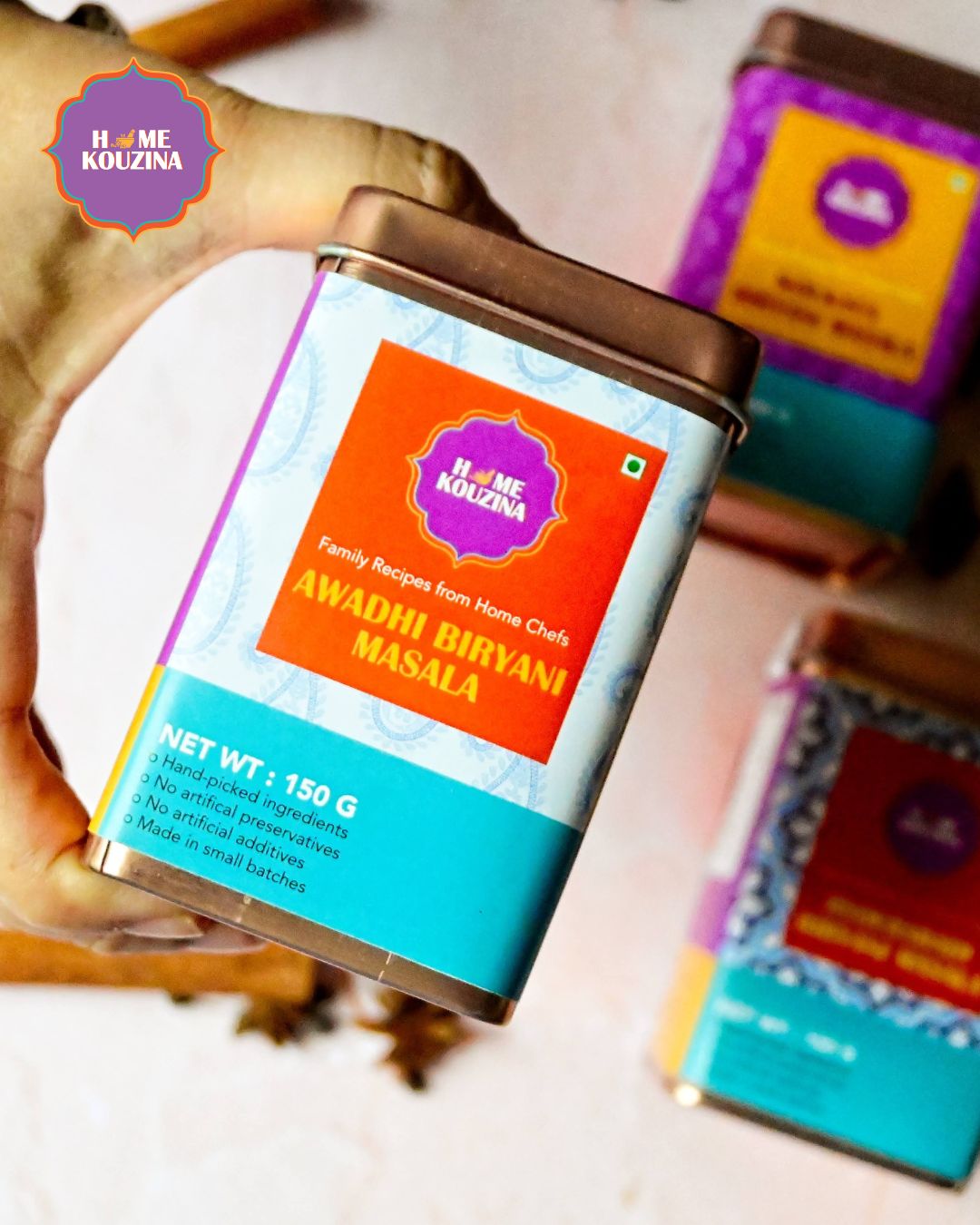 Hand holding Home Kouzina Awadhi Biryani Masala tin from Biryanis of India Premium Tin Pack – whole spices scattered in the background – authentic Lucknow biryani flavor