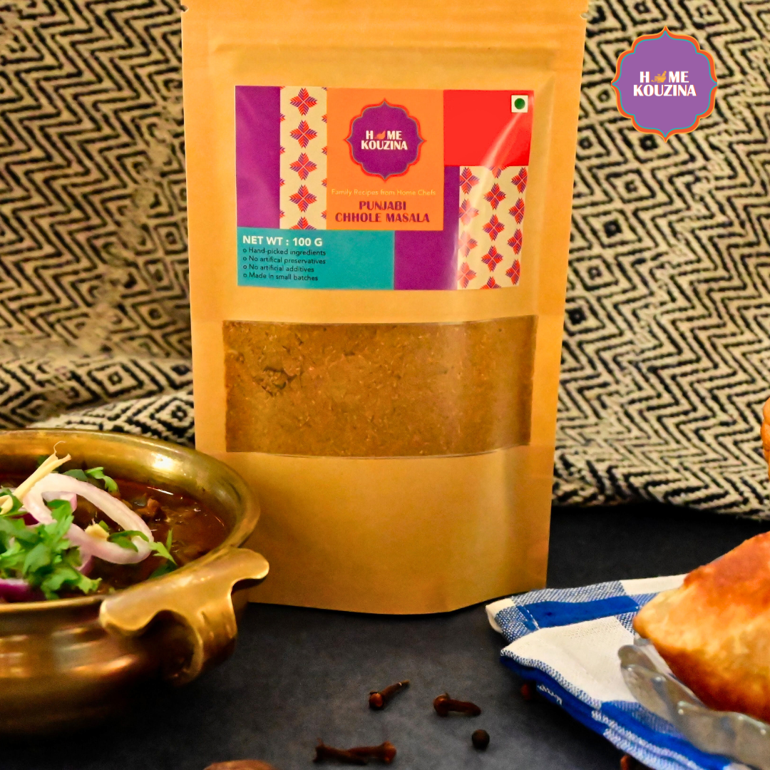 Front view of Home Kouzina Punjabi Chhole Masala 100g pouch on table with punjabi chhole in a brass bowl garnished with onions and coriander wiht kulche in a plate – traditional, all-natural Punjabi curry masala