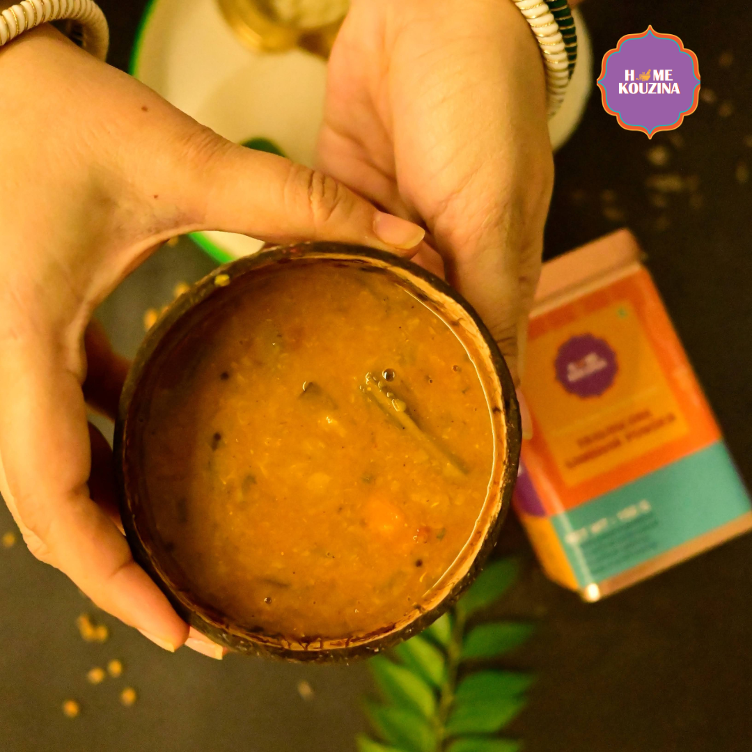 Top view of bowl of sambhar held in hand with Home Kouzina Travancore Sambhar Powder 150g tin on table – small-batch, all-natural Kerala sambhar masala