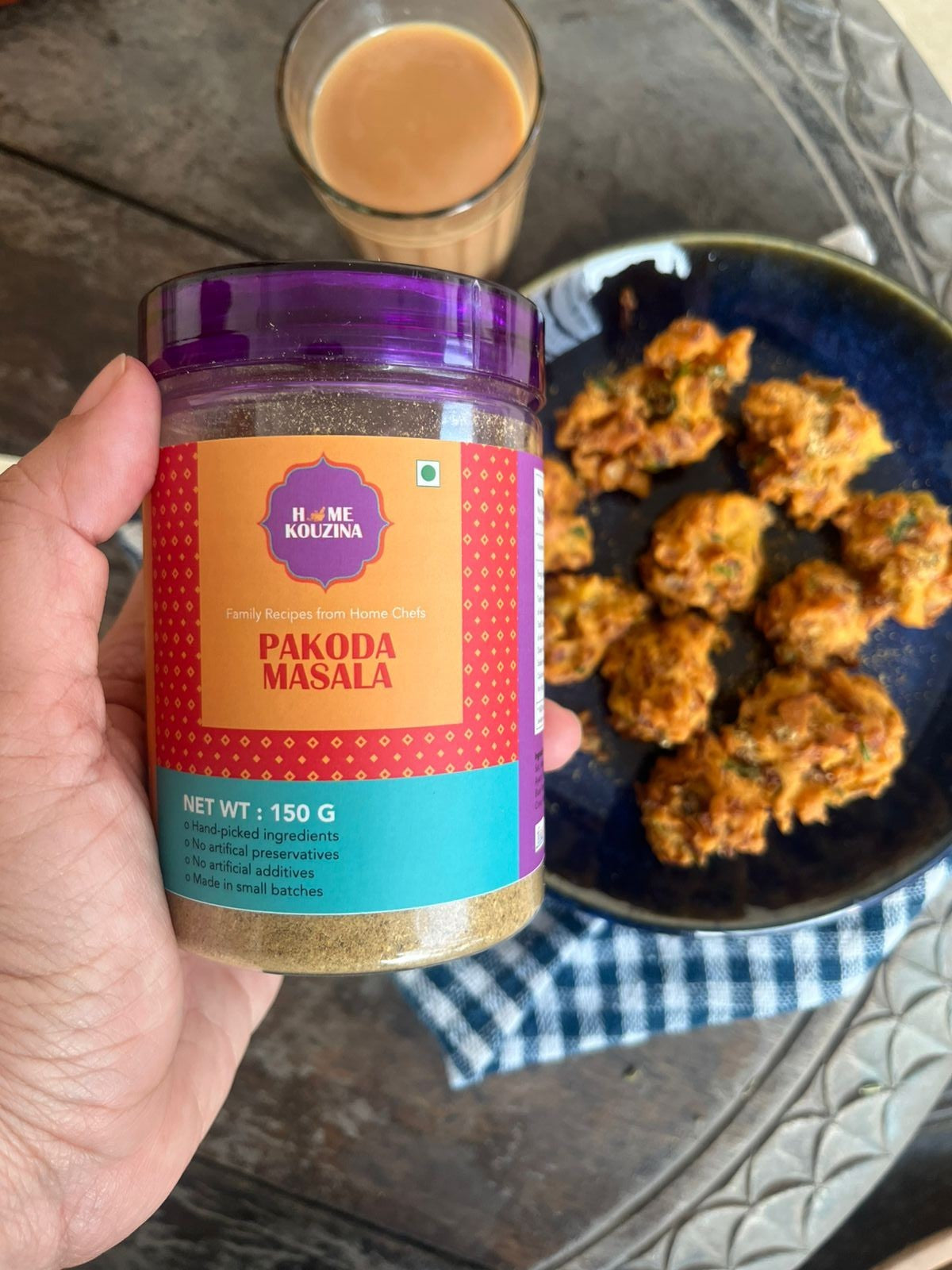Hand holding a jar of 'Home kouzina' Pakoda Masala with fried pakodas and a glass of drink on a table.