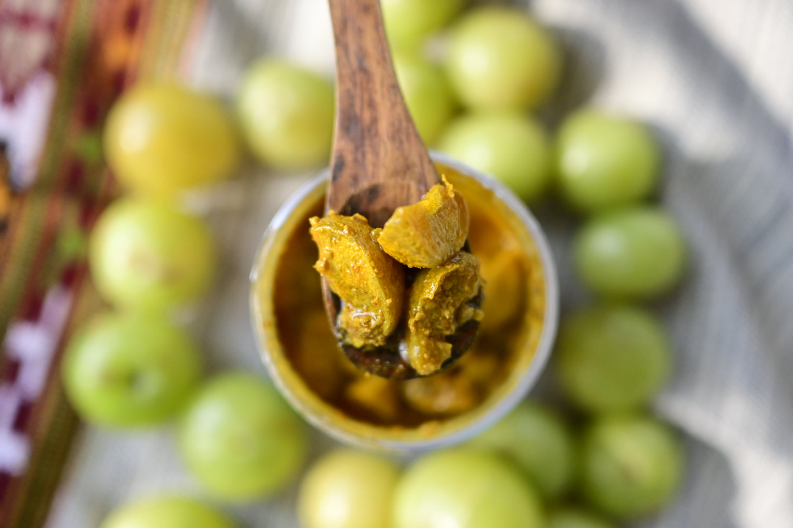 Spoonful of Amla Achaar pickle in a container surrounded by amla or gooseberry fruits.