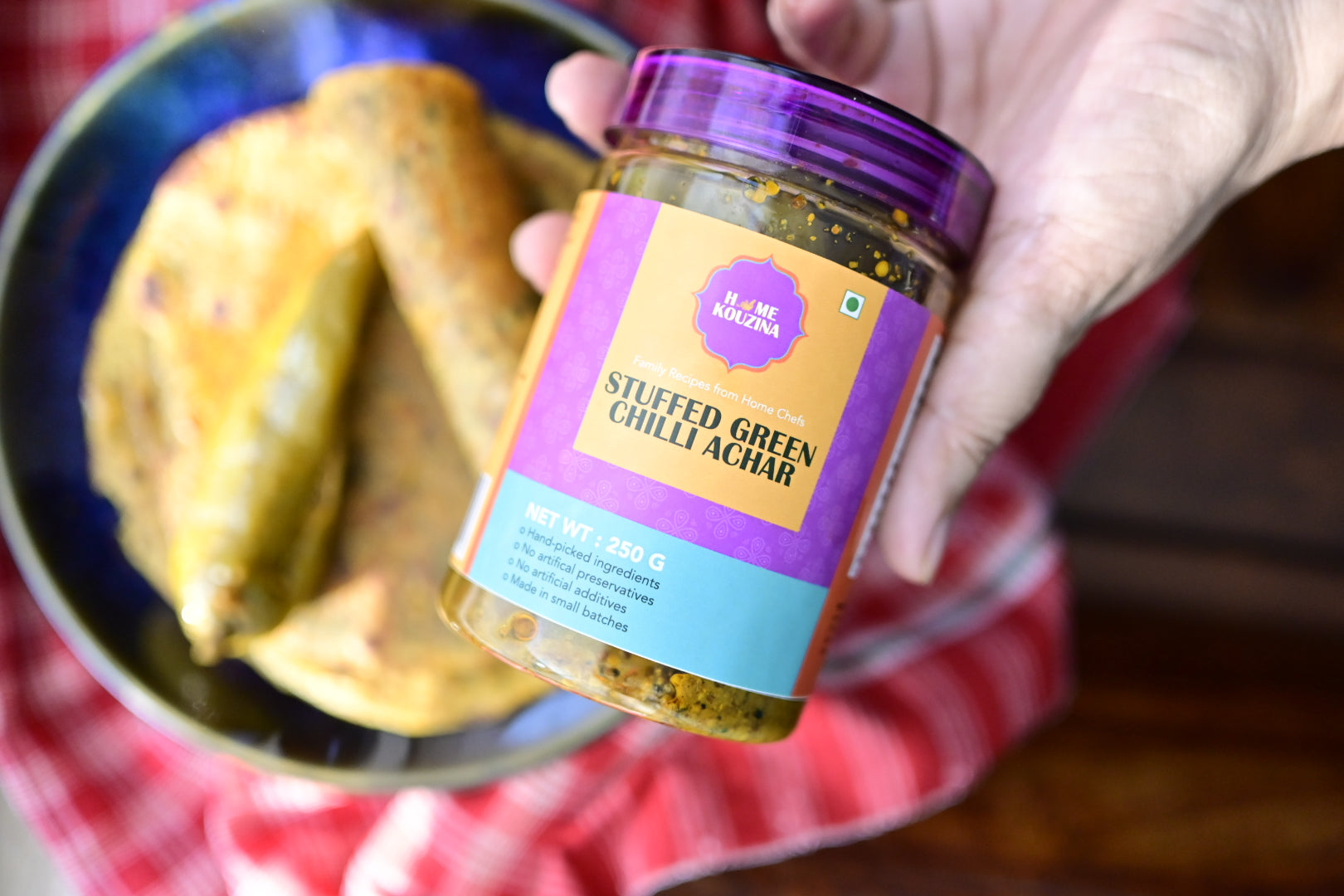 Jar of 'Stuffed Green Chilli Achar' held by a hand with food in the background