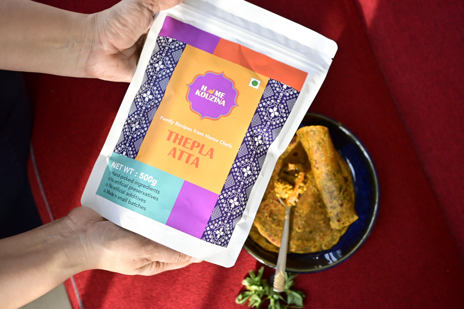 Hand holding a packet of 'Thepla Aita' with a plate of thepla in the background on a red fabric.